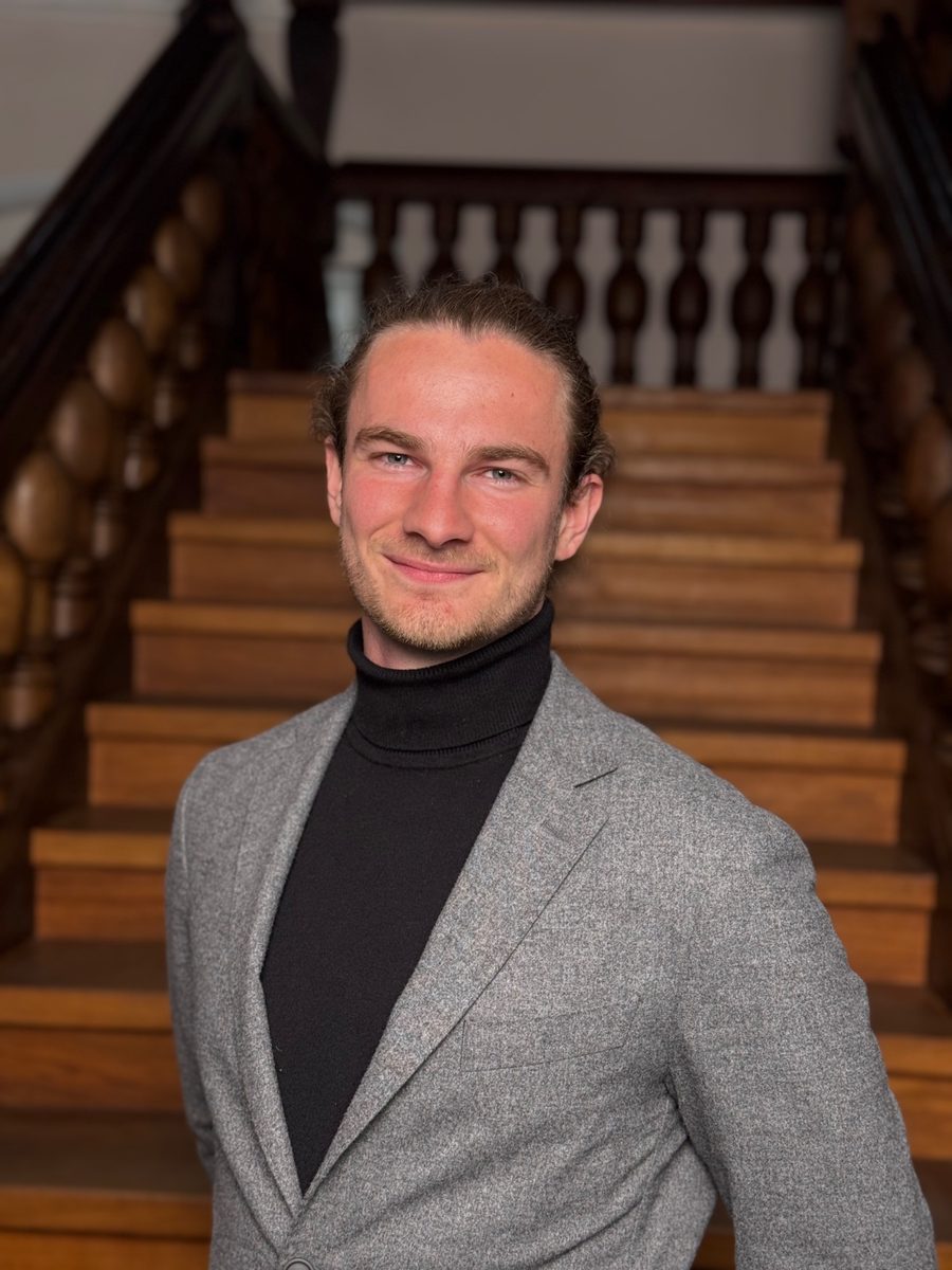 Maximilian Pfannkuch mit Rollkragenpullover und grauem Sakko vor einer Holztreppe